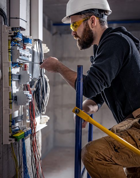 A male electrician works in a switchboard with an electrical connecting cable, connects the equipment with tools.