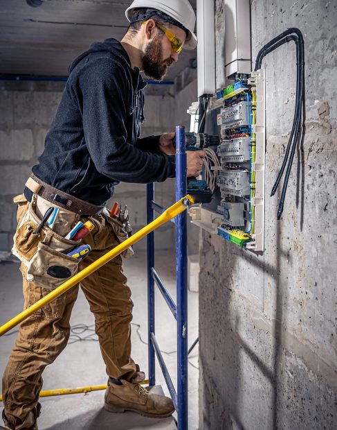 A male electrician works in a switchboard with an electrical connecting cable, connects the equipment with tools.